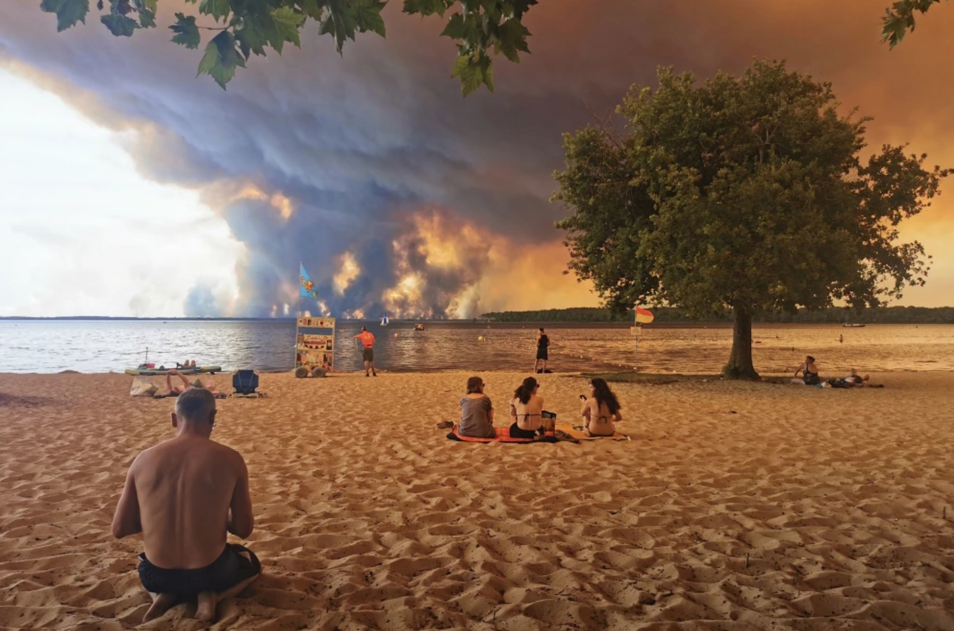 People sitting on a beach watching a massive wildfire burn across the lake